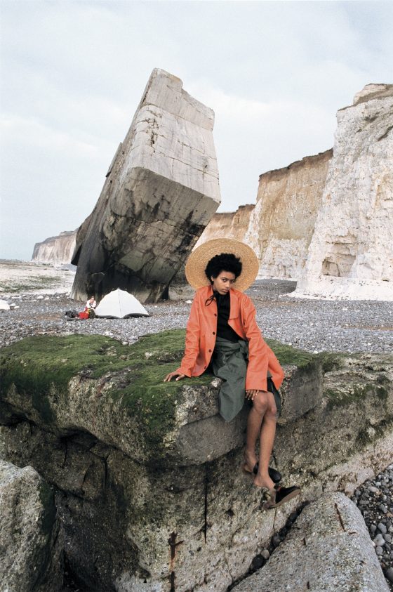 Coat, pleated top & skirt by Jil Sander, parka around the waist from stylist’s archive, oversized brimmed hat by Jacquemus, moccasins by Gucci.